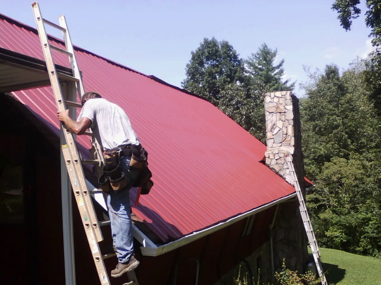 Roofer on a ladder beside a red metal roof and stone chimney — Soffit & Fascia Repair by Roofing Brothers in Hughesville